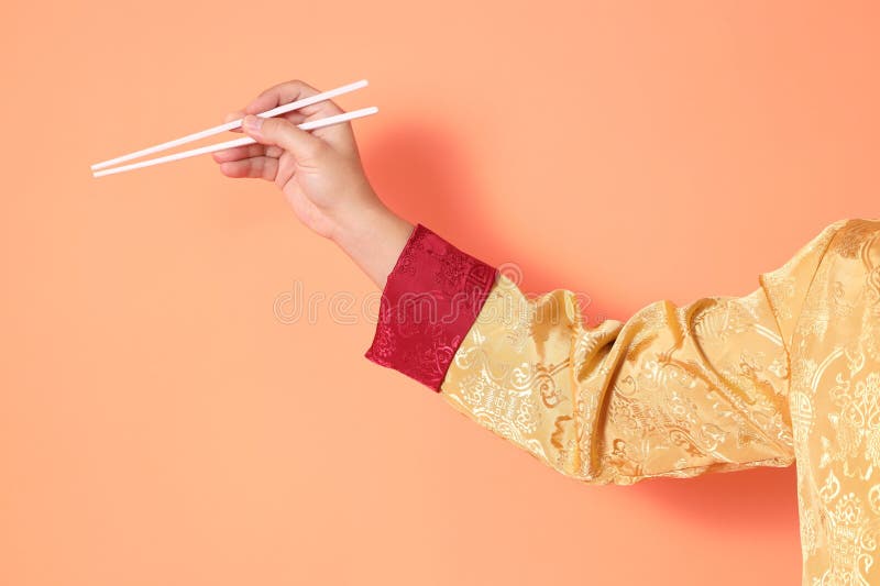 Happy Chinese new year. asian chinese senior man wearing golden traditional cheongsam qipao or changshan dress with gesture of hand holding white chopsticks isolated on orange background. Happy Chinese new year. asian chinese senior man wearing golden traditional cheongsam qipao or changshan dress with gesture of hand holding white chopsticks isolated on orange background