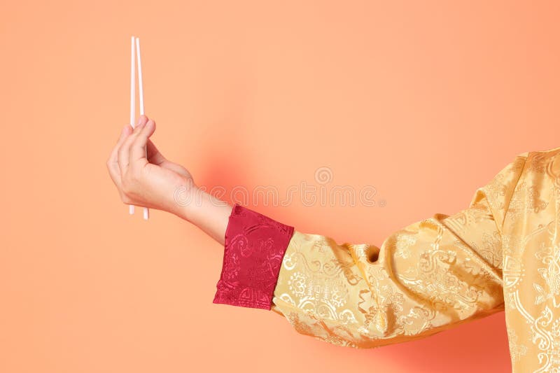 Happy Chinese new year. asian chinese senior man wearing golden traditional cheongsam qipao or changshan dress with gesture of hand holding white chopsticks isolated on orange background. Happy Chinese new year. asian chinese senior man wearing golden traditional cheongsam qipao or changshan dress with gesture of hand holding white chopsticks isolated on orange background