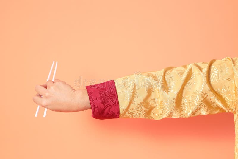 Happy Chinese new year. asian chinese senior man wearing golden traditional cheongsam qipao or changshan dress with gesture of hand holding white chopsticks isolated on orange background. Happy Chinese new year. asian chinese senior man wearing golden traditional cheongsam qipao or changshan dress with gesture of hand holding white chopsticks isolated on orange background