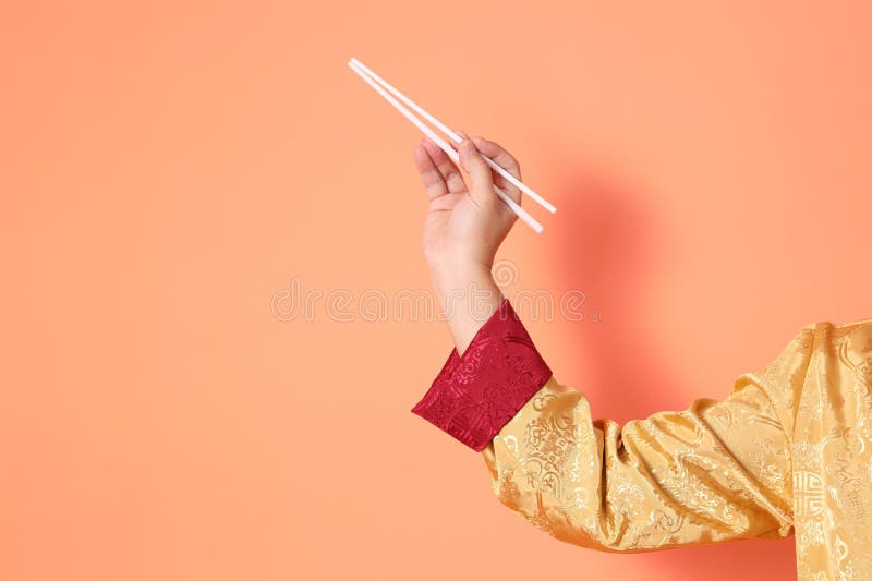 Happy Chinese new year. asian chinese senior man wearing golden traditional cheongsam qipao or changshan dress with gesture of hand holding white chopsticks isolated on orange background. Happy Chinese new year. asian chinese senior man wearing golden traditional cheongsam qipao or changshan dress with gesture of hand holding white chopsticks isolated on orange background