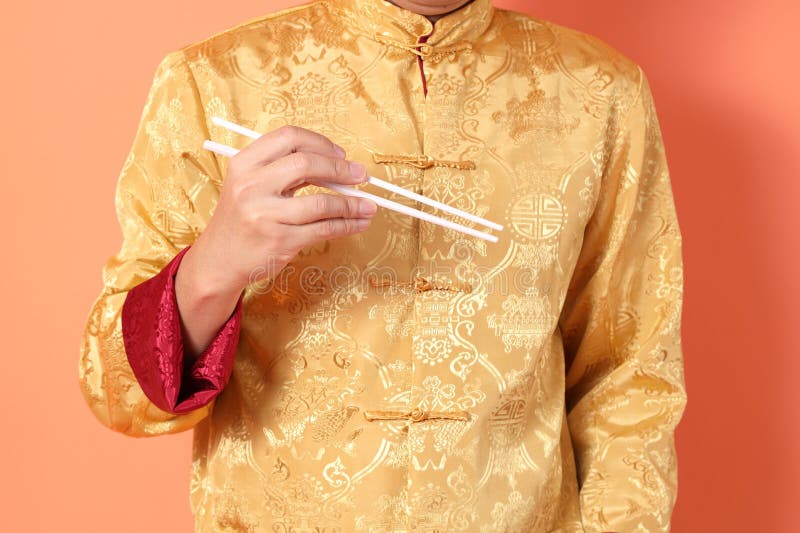 Happy Chinese new year. asian chinese senior man wearing golden traditional cheongsam qipao or changshan dress with gesture of hand holding white chopsticks isolated on orange background. Happy Chinese new year. asian chinese senior man wearing golden traditional cheongsam qipao or changshan dress with gesture of hand holding white chopsticks isolated on orange background