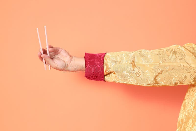 Happy Chinese new year. asian chinese senior man wearing golden traditional cheongsam qipao or changshan dress with gesture of hand holding white chopsticks isolated on orange background. Happy Chinese new year. asian chinese senior man wearing golden traditional cheongsam qipao or changshan dress with gesture of hand holding white chopsticks isolated on orange background