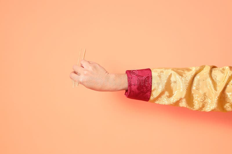 Happy Chinese new year. asian chinese senior man wearing golden traditional cheongsam qipao or changshan dress with gesture of hand holding wood chopsticks isolated on orange background. Happy Chinese new year. asian chinese senior man wearing golden traditional cheongsam qipao or changshan dress with gesture of hand holding wood chopsticks isolated on orange background