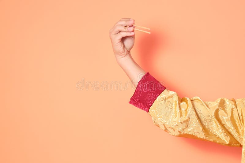 Happy Chinese new year. asian chinese senior man wearing golden traditional cheongsam qipao or changshan dress with gesture of hand holding wood chopsticks isolated on orange background. Happy Chinese new year. asian chinese senior man wearing golden traditional cheongsam qipao or changshan dress with gesture of hand holding wood chopsticks isolated on orange background