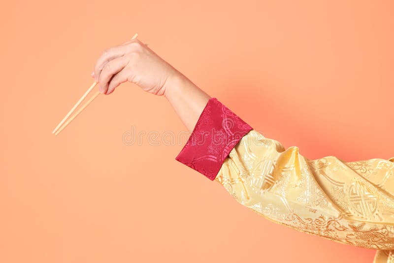 Happy Chinese new year. asian chinese senior man wearing golden traditional cheongsam qipao or changshan dress with gesture of hand holding wood chopsticks isolated on orange background. Happy Chinese new year. asian chinese senior man wearing golden traditional cheongsam qipao or changshan dress with gesture of hand holding wood chopsticks isolated on orange background