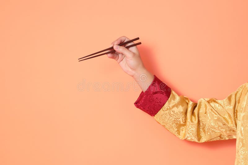 Happy Chinese new year. asian chinese senior man wearing golden traditional cheongsam qipao or changshan dress with gesture of hand holding brown chopsticks isolated on orange background. Happy Chinese new year. asian chinese senior man wearing golden traditional cheongsam qipao or changshan dress with gesture of hand holding brown chopsticks isolated on orange background