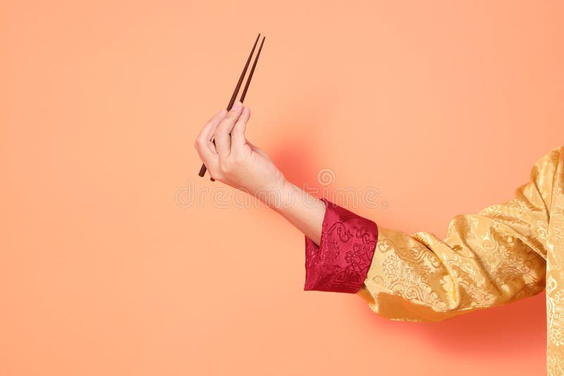 Happy Chinese new year. asian chinese senior man wearing golden traditional cheongsam qipao or changshan dress with gesture of hand holding brown chopsticks isolated on orange background. Happy Chinese new year. asian chinese senior man wearing golden traditional cheongsam qipao or changshan dress with gesture of hand holding brown chopsticks isolated on orange background