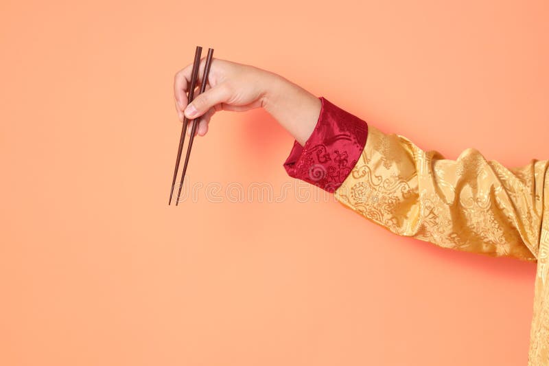 Happy Chinese new year. asian chinese senior man wearing golden traditional cheongsam qipao or changshan dress with gesture of hand holding brown chopsticks isolated on orange background. Happy Chinese new year. asian chinese senior man wearing golden traditional cheongsam qipao or changshan dress with gesture of hand holding brown chopsticks isolated on orange background