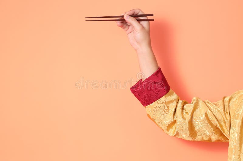 Happy Chinese new year. asian chinese senior man wearing golden traditional cheongsam qipao or changshan dress with gesture of hand holding brown chopsticks isolated on orange background. Happy Chinese new year. asian chinese senior man wearing golden traditional cheongsam qipao or changshan dress with gesture of hand holding brown chopsticks isolated on orange background