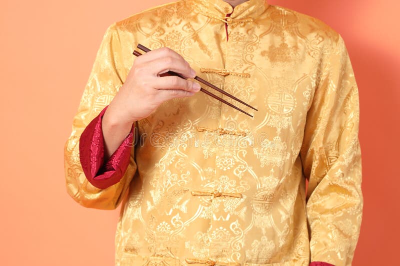 Happy Chinese new year. asian chinese senior man wearing golden traditional cheongsam qipao or changshan dress with gesture of hand holding brown chopsticks isolated on orange background. Happy Chinese new year. asian chinese senior man wearing golden traditional cheongsam qipao or changshan dress with gesture of hand holding brown chopsticks isolated on orange background