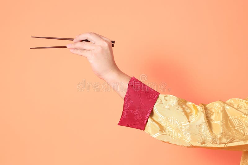 Happy Chinese new year. asian chinese senior man wearing golden traditional cheongsam qipao or changshan dress with gesture of hand holding brown chopsticks isolated on orange background. Happy Chinese new year. asian chinese senior man wearing golden traditional cheongsam qipao or changshan dress with gesture of hand holding brown chopsticks isolated on orange background