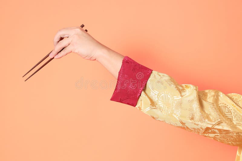 Happy Chinese new year. asian chinese senior man wearing golden traditional cheongsam qipao or changshan dress with gesture of hand holding brown chopsticks isolated on orange background. Happy Chinese new year. asian chinese senior man wearing golden traditional cheongsam qipao or changshan dress with gesture of hand holding brown chopsticks isolated on orange background