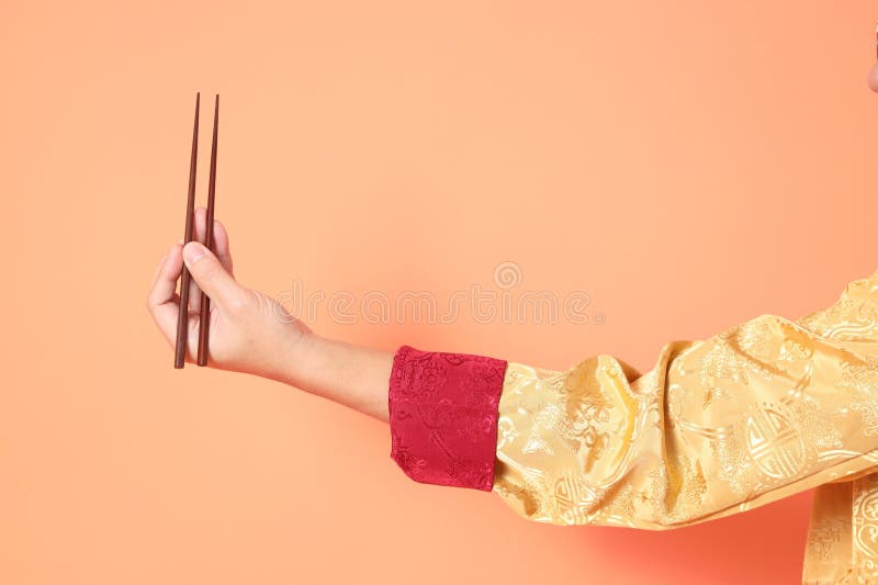 Happy Chinese new year. asian chinese senior man wearing golden traditional cheongsam qipao or changshan dress with gesture of hand holding brown chopsticks isolated on orange background. Happy Chinese new year. asian chinese senior man wearing golden traditional cheongsam qipao or changshan dress with gesture of hand holding brown chopsticks isolated on orange background