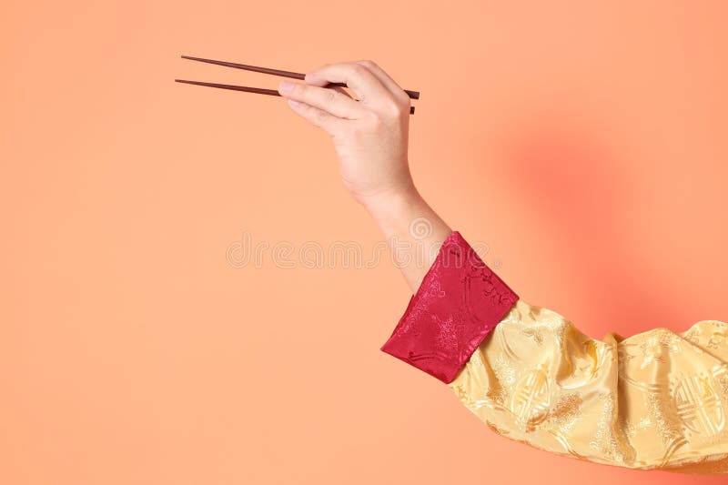 Happy Chinese new year. asian chinese senior man wearing golden traditional cheongsam qipao or changshan dress with gesture of hand holding brown chopsticks isolated on orange background. Happy Chinese new year. asian chinese senior man wearing golden traditional cheongsam qipao or changshan dress with gesture of hand holding brown chopsticks isolated on orange background