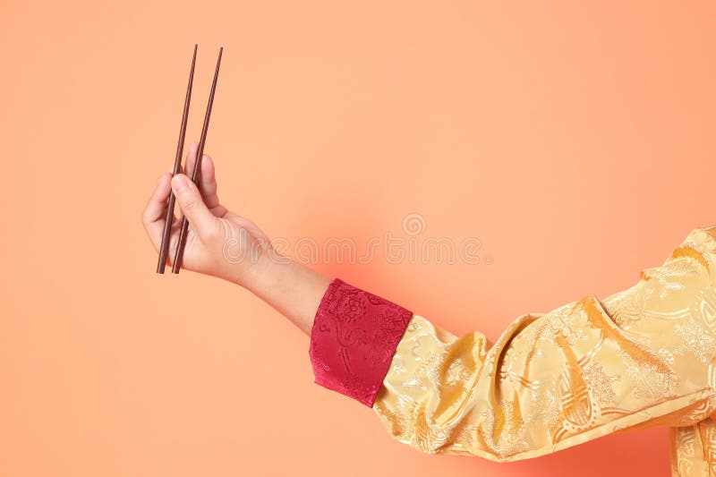 Happy Chinese new year. asian chinese senior man wearing golden traditional cheongsam qipao or changshan dress with gesture of hand holding silver chopsticks isolated on orange background. Happy Chinese new year. asian chinese senior man wearing golden traditional cheongsam qipao or changshan dress with gesture of hand holding silver chopsticks isolated on orange background