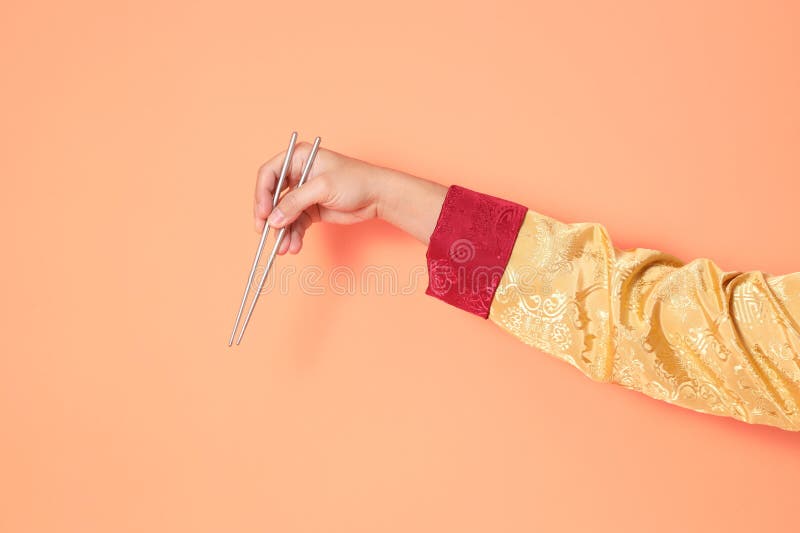 Happy Chinese new year. asian chinese senior man wearing golden traditional cheongsam qipao or changshan dress with gesture of hand holding silver chopsticks isolated on orange background. Happy Chinese new year. asian chinese senior man wearing golden traditional cheongsam qipao or changshan dress with gesture of hand holding silver chopsticks isolated on orange background