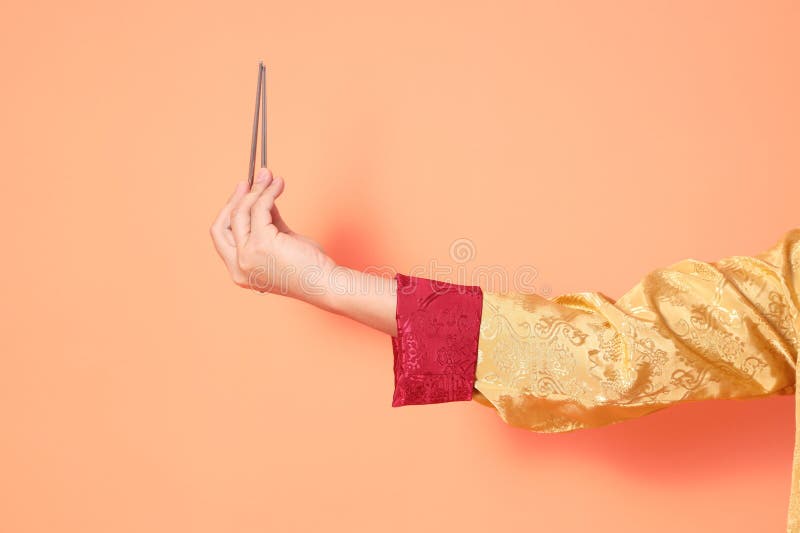 Happy Chinese new year. asian chinese senior man wearing golden traditional cheongsam qipao or changshan dress with gesture of hand holding silver chopsticks isolated on orange background. Happy Chinese new year. asian chinese senior man wearing golden traditional cheongsam qipao or changshan dress with gesture of hand holding silver chopsticks isolated on orange background