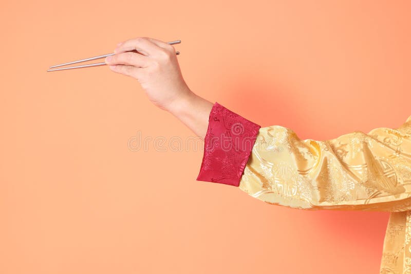 Happy Chinese new year. asian chinese senior man wearing golden traditional cheongsam qipao or changshan dress with gesture of hand holding silver chopsticks isolated on orange background. Happy Chinese new year. asian chinese senior man wearing golden traditional cheongsam qipao or changshan dress with gesture of hand holding silver chopsticks isolated on orange background