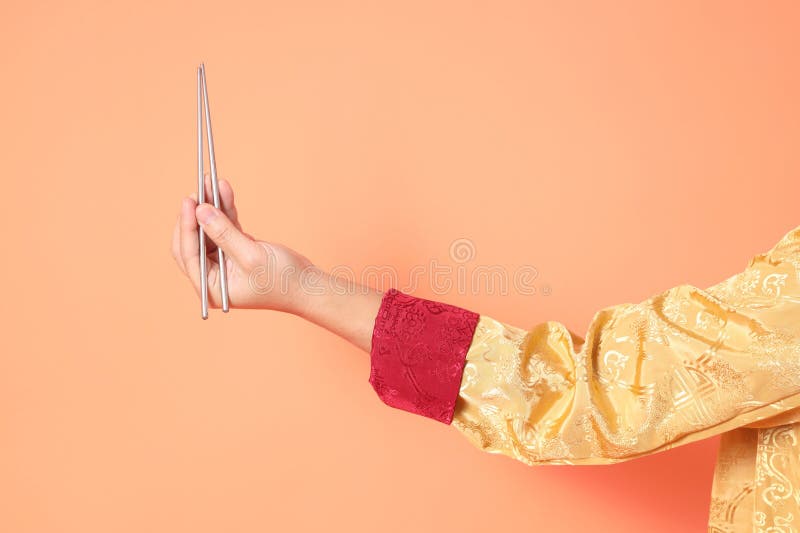 Happy Chinese new year. asian chinese senior man wearing golden traditional cheongsam qipao or changshan dress with gesture of hand holding silver chopsticks isolated on orange background. Happy Chinese new year. asian chinese senior man wearing golden traditional cheongsam qipao or changshan dress with gesture of hand holding silver chopsticks isolated on orange background