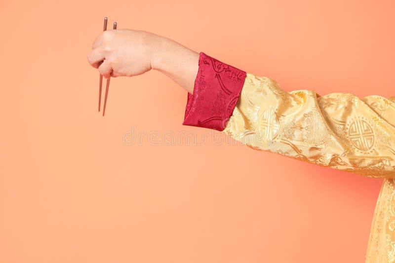 Happy Chinese new year. asian chinese senior man wearing golden traditional cheongsam qipao or changshan dress with gesture of hand holding silver chopsticks isolated on orange background. Happy Chinese new year. asian chinese senior man wearing golden traditional cheongsam qipao or changshan dress with gesture of hand holding silver chopsticks isolated on orange background