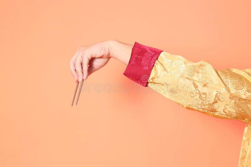 Happy Chinese new year. asian chinese senior man wearing golden traditional cheongsam qipao or changshan dress with gesture of hand holding silver chopsticks isolated on orange background. Happy Chinese new year. asian chinese senior man wearing golden traditional cheongsam qipao or changshan dress with gesture of hand holding silver chopsticks isolated on orange background