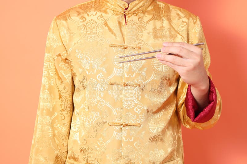 Happy Chinese new year. asian chinese senior man wearing golden traditional cheongsam qipao or changshan dress with gesture of hand holding silver chopsticks isolated on orange background. Happy Chinese new year. asian chinese senior man wearing golden traditional cheongsam qipao or changshan dress with gesture of hand holding silver chopsticks isolated on orange background