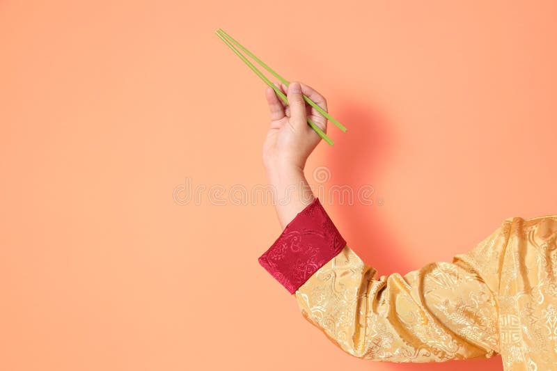 Happy Chinese new year. asian chinese senior man wearing golden traditional cheongsam qipao or changshan dress with gesture of hand holding green chopsticks isolated on orange background. Happy Chinese new year. asian chinese senior man wearing golden traditional cheongsam qipao or changshan dress with gesture of hand holding green chopsticks isolated on orange background