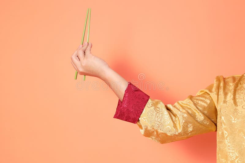Happy Chinese new year. asian chinese senior man wearing golden traditional cheongsam qipao or changshan dress with gesture of hand holding green chopsticks isolated on orange background. Happy Chinese new year. asian chinese senior man wearing golden traditional cheongsam qipao or changshan dress with gesture of hand holding green chopsticks isolated on orange background