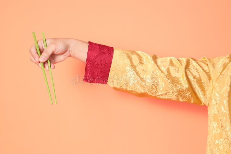 Happy Chinese new year. asian chinese senior man wearing golden traditional cheongsam qipao or changshan dress with gesture of hand holding green chopsticks isolated on orange background. Happy Chinese new year. asian chinese senior man wearing golden traditional cheongsam qipao or changshan dress with gesture of hand holding green chopsticks isolated on orange background