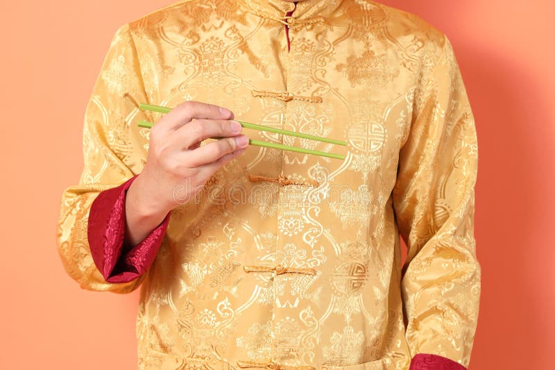 Happy Chinese new year. asian chinese senior man wearing golden traditional cheongsam qipao or changshan dress with gesture of hand holding green chopsticks isolated on orange background. Happy Chinese new year. asian chinese senior man wearing golden traditional cheongsam qipao or changshan dress with gesture of hand holding green chopsticks isolated on orange background