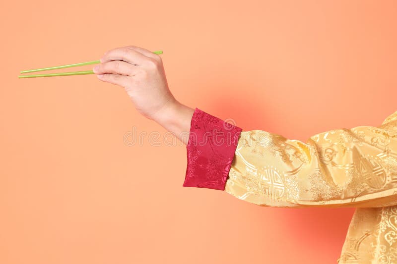 Happy Chinese new year. asian chinese senior man wearing golden traditional cheongsam qipao or changshan dress with gesture of hand holding green chopsticks isolated on orange background. Happy Chinese new year. asian chinese senior man wearing golden traditional cheongsam qipao or changshan dress with gesture of hand holding green chopsticks isolated on orange background