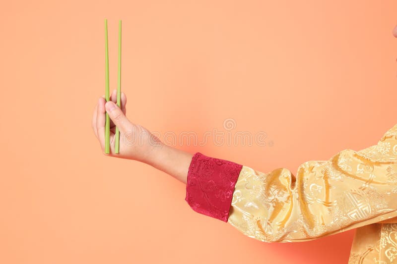 Happy Chinese new year. asian chinese senior man wearing golden traditional cheongsam qipao or changshan dress with gesture of hand holding green chopsticks isolated on orange background. Happy Chinese new year. asian chinese senior man wearing golden traditional cheongsam qipao or changshan dress with gesture of hand holding green chopsticks isolated on orange background