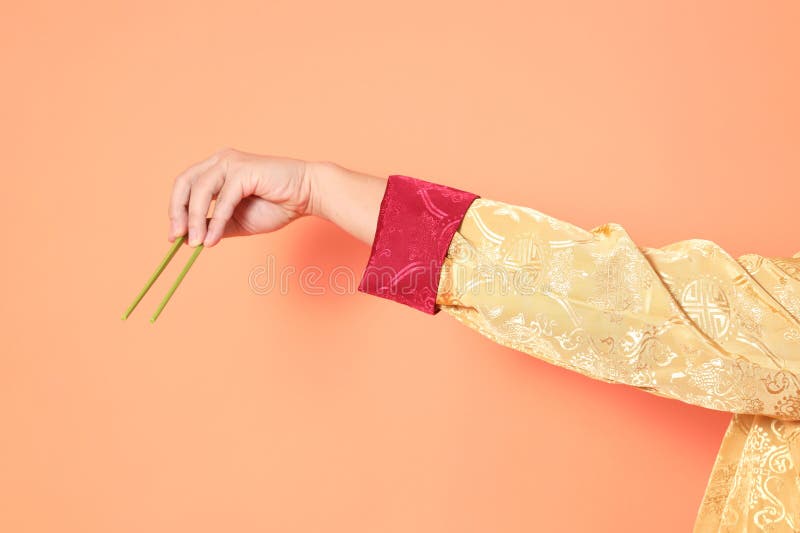 Happy Chinese new year. asian chinese senior man wearing golden traditional cheongsam qipao or changshan dress with gesture of hand holding green chopsticks isolated on orange background. Happy Chinese new year. asian chinese senior man wearing golden traditional cheongsam qipao or changshan dress with gesture of hand holding green chopsticks isolated on orange background