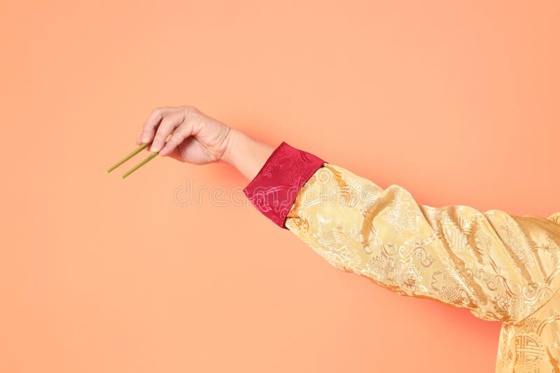 Happy Chinese new year. asian chinese senior man wearing golden traditional cheongsam qipao or changshan dress with gesture of hand holding green chopsticks isolated on orange background. Happy Chinese new year. asian chinese senior man wearing golden traditional cheongsam qipao or changshan dress with gesture of hand holding green chopsticks isolated on orange background