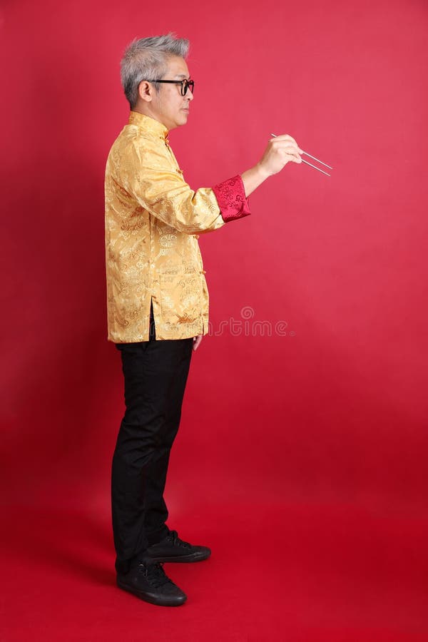 Happy Chinese new year. Asian Chinese energetic senior man wearing golden traditional cheongsam qipao or changshan dress with gesture of hand holding silver chopsticks isolated on red background. Happy Chinese new year. Asian Chinese energetic senior man wearing golden traditional cheongsam qipao or changshan dress with gesture of hand holding silver chopsticks isolated on red background