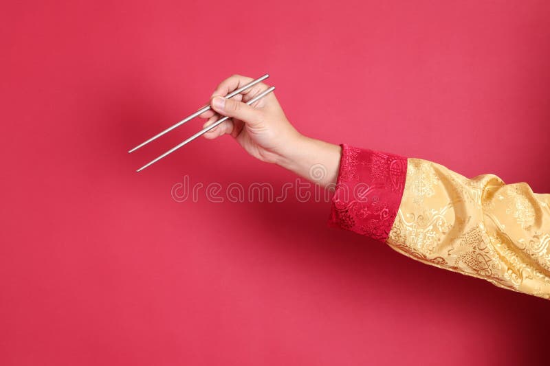 Happy Chinese new year. Asian Chinese energetic senior man wearing golden traditional cheongsam qipao or changshan dress with gesture of hand holding silver chopsticks isolated on red background. Happy Chinese new year. Asian Chinese energetic senior man wearing golden traditional cheongsam qipao or changshan dress with gesture of hand holding silver chopsticks isolated on red background