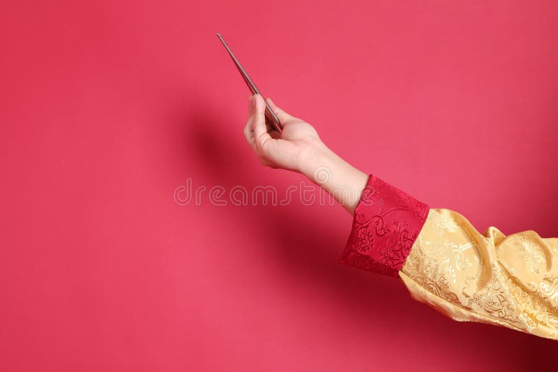 Happy Chinese new year. Asian Chinese energetic senior man wearing golden traditional cheongsam qipao or changshan dress with gesture of hand holding silver chopsticks isolated on red background. Happy Chinese new year. Asian Chinese energetic senior man wearing golden traditional cheongsam qipao or changshan dress with gesture of hand holding silver chopsticks isolated on red background