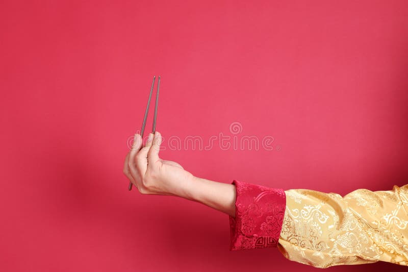 Happy Chinese new year. Asian Chinese energetic senior man wearing golden traditional cheongsam qipao or changshan dress with gesture of hand holding silver chopsticks isolated on red background. Happy Chinese new year. Asian Chinese energetic senior man wearing golden traditional cheongsam qipao or changshan dress with gesture of hand holding silver chopsticks isolated on red background