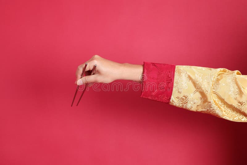 Happy Chinese new year. Asian Chinese energetic senior man wearing golden traditional cheongsam qipao or changshan dress with gesture of hand holding silver chopsticks isolated on red background. Happy Chinese new year. Asian Chinese energetic senior man wearing golden traditional cheongsam qipao or changshan dress with gesture of hand holding silver chopsticks isolated on red background