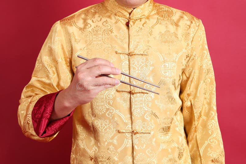 Happy Chinese new year. Asian Chinese energetic senior man wearing golden traditional cheongsam qipao or changshan dress with gesture of hand holding silver chopsticks isolated on red background. Happy Chinese new year. Asian Chinese energetic senior man wearing golden traditional cheongsam qipao or changshan dress with gesture of hand holding silver chopsticks isolated on red background