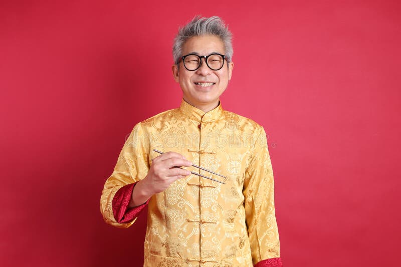 Happy Chinese new year. Asian Chinese energetic senior man wearing golden traditional cheongsam qipao or changshan dress with gesture of hand holding silver chopsticks isolated on red background. Happy Chinese new year. Asian Chinese energetic senior man wearing golden traditional cheongsam qipao or changshan dress with gesture of hand holding silver chopsticks isolated on red background