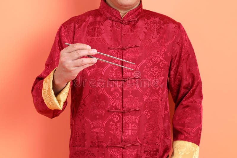 Happy Chinese new year. Hand of asian chinese senior man wearing red traditional cheongsam qipao or changshan dress with gesture of hand holding silver chopsticks isolated on orange background. Happy Chinese new year. Hand of asian chinese senior man wearing red traditional cheongsam qipao or changshan dress with gesture of hand holding silver chopsticks isolated on orange background