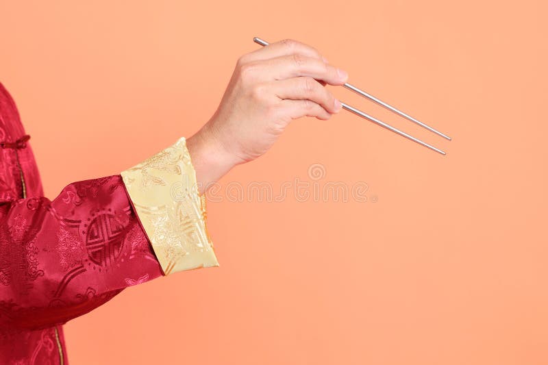 Happy Chinese new year. Hand of asian chinese senior man wearing red traditional cheongsam qipao or changshan dress with gesture of hand holding silver chopsticks isolated on orange background. Happy Chinese new year. Hand of asian chinese senior man wearing red traditional cheongsam qipao or changshan dress with gesture of hand holding silver chopsticks isolated on orange background