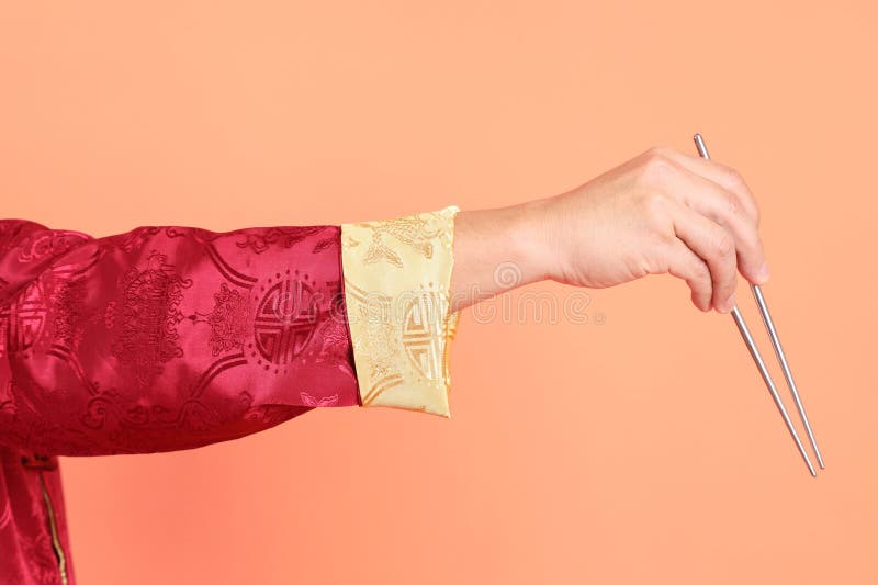 Happy Chinese new year. Hand of asian chinese senior man wearing red traditional cheongsam qipao or changshan dress with gesture of hand holding silver chopsticks isolated on orange background. Happy Chinese new year. Hand of asian chinese senior man wearing red traditional cheongsam qipao or changshan dress with gesture of hand holding silver chopsticks isolated on orange background