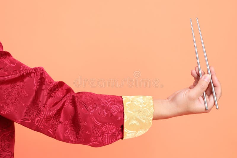 Happy Chinese new year. Hand of asian chinese senior man wearing red traditional cheongsam qipao or changshan dress with gesture of hand holding silver chopsticks isolated on orange background. Happy Chinese new year. Hand of asian chinese senior man wearing red traditional cheongsam qipao or changshan dress with gesture of hand holding silver chopsticks isolated on orange background