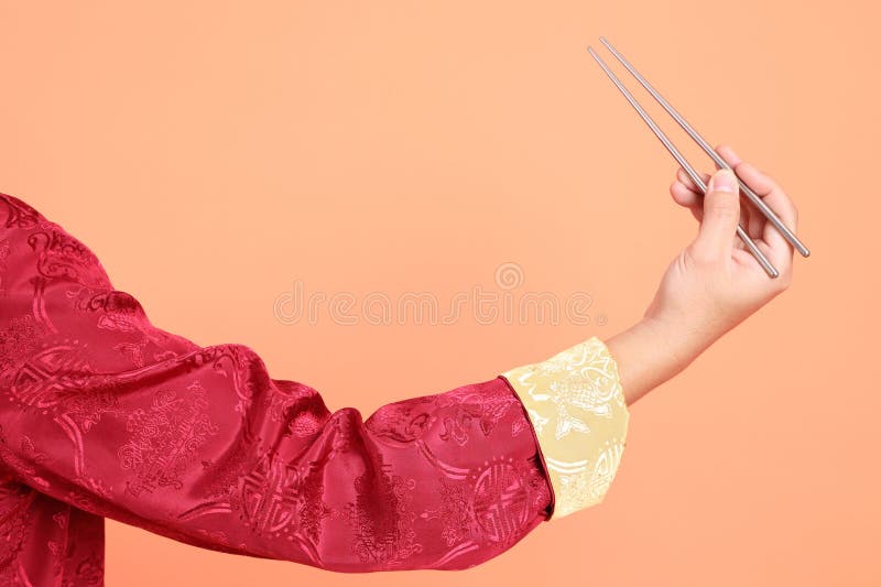 Happy Chinese new year. Hand of asian chinese senior man wearing red traditional cheongsam qipao or changshan dress with gesture of hand holding silver chopsticks isolated on orange background. Happy Chinese new year. Hand of asian chinese senior man wearing red traditional cheongsam qipao or changshan dress with gesture of hand holding silver chopsticks isolated on orange background