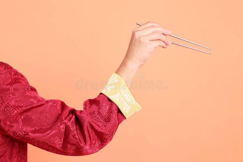 Happy Chinese new year. Hand of asian chinese senior man wearing red traditional cheongsam qipao or changshan dress with gesture of hand holding silver chopsticks isolated on orange background. Happy Chinese new year. Hand of asian chinese senior man wearing red traditional cheongsam qipao or changshan dress with gesture of hand holding silver chopsticks isolated on orange background