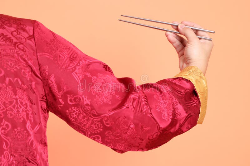 Happy Chinese new year. Hand of asian chinese senior man wearing red traditional cheongsam qipao or changshan dress with gesture of hand holding silver chopsticks isolated on orange background. Happy Chinese new year. Hand of asian chinese senior man wearing red traditional cheongsam qipao or changshan dress with gesture of hand holding silver chopsticks isolated on orange background