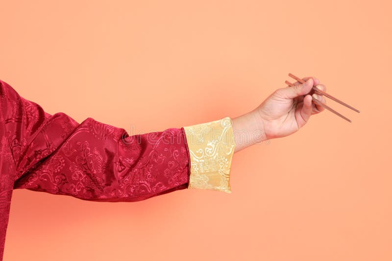 Happy Chinese new year. Hand of asian chinese senior man wearing red traditional cheongsam qipao or changshan dress with gesture of hand holding silver chopsticks isolated on orange background. Happy Chinese new year. Hand of asian chinese senior man wearing red traditional cheongsam qipao or changshan dress with gesture of hand holding silver chopsticks isolated on orange background