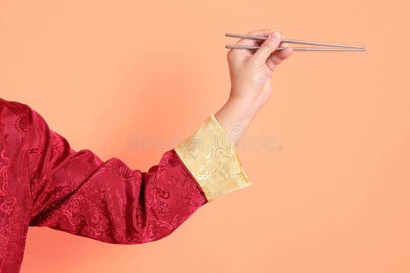 Happy Chinese new year. Hand of asian chinese senior man wearing red traditional cheongsam qipao or changshan dress with gesture of hand holding silver chopsticks isolated on orange background. Happy Chinese new year. Hand of asian chinese senior man wearing red traditional cheongsam qipao or changshan dress with gesture of hand holding silver chopsticks isolated on orange background